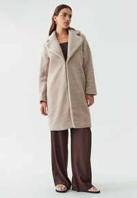 Abrigo de forro polar beige con cuello chal, mangas largas y corte recto; llevado sobre pantalones de pierna ancha marrones y sandalias claras.