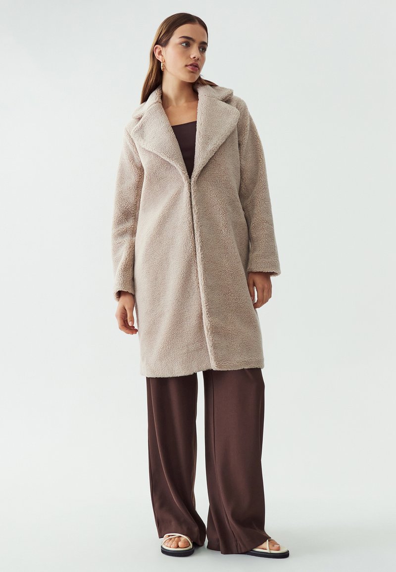 Abrigo de forro polar beige con cuello chal, mangas largas y corte recto; llevado sobre pantalones de pierna ancha marrones y sandalias claras.