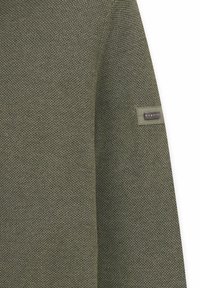 Olivgrüner gestrickter Pullover mit strukturierter Oberfläche und einem Etikett am Ärmel. Der Stoff ist weich und weist ein dezentes Webmuster auf.