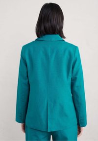 Marynarka w kolorze morskim, o dopasowanym kroju, z wyciętym kołnierzem, pojedynczym rozcięciem z tyłu i mankietami na guziki. Wykonana z gładkiej tkaniny, prezentująca czysty, schludny design.