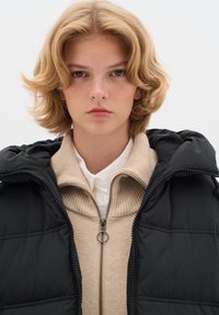 Svart pufferväst med huva, lagd över en beige ribbad zip-up tröja, med en vit skjorta under. Slät textur.
