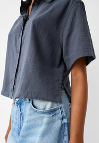 Koszula na krótki rękaw zapinana na guziki w ciemnoszarym materiale o teksturowanym wykończeniu. Posiada skrócony krój oraz boczne rozcięcia. W połączeniu z jasnoniebieskimi dżinsami.
