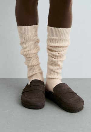 Legs wearing beige slouchy knit leg warmers over brown leggings with dark brown suede slip-on loafers on a gray floor.