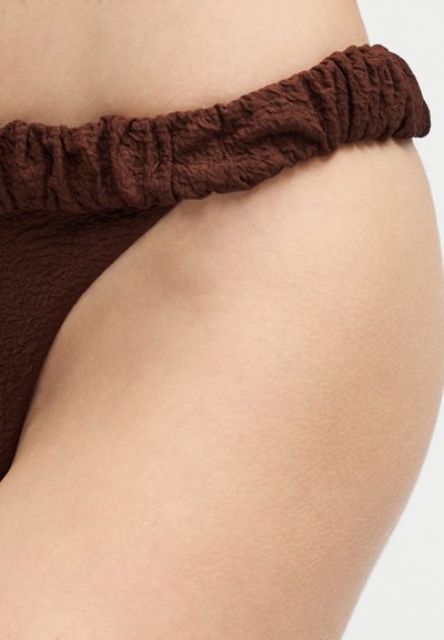 Gros plan sur la hanche d'une personne portant un bas de bikini texturé marron foncé avec une taille élastique froncée, sur un fond uni.