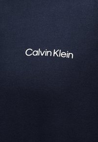 Σκούρο μπλε ύφασμα με το λευκό λογότυπο Calvin Klein στο κέντρο, σε απλή, μοντέρνα γραμματοσειρά.