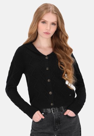 Ung kvinde med langt bølget blondt hår iført en sort knappet cardigan og mørke jeans med hænderne i lommerne mod en ensfarvet baggrund.