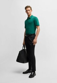 Polo verde con maniche corte, pantaloni neri su misura e scarpe nere. Porta una borsa nera in pelle. Design semplice e moderno.
