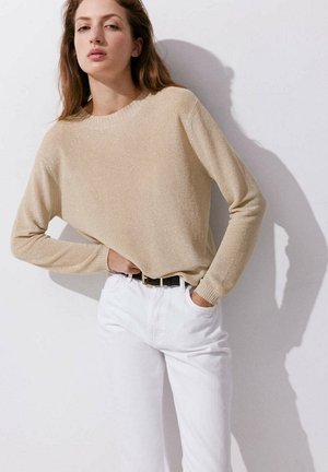 Jeune femme portant un pull beige à manches longues et un pantalon blanc, debout avec une main dans la poche contre un mur blanc.