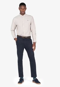 Camisa de botones color beige claro, chinos azul marino, cinturón trenzado y zapatillas oscuras. Diseño sencillo, ajuste a medida y acentos mínimos.