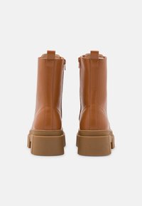 Botas de tornozelo em imitação de couro castanho com sola grossa, fecho de zipper na parte traseira e detalhes contrastantes em tons mais claros. Textura suave, costura mínima.