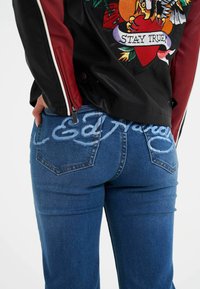 Sort læderjakke med røde ærmer, prydet med farverig broderi. Blå denim jeans med lyseblåt skrift på taljen.