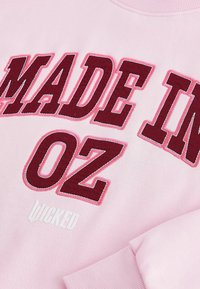 Růžová mikina s výrazným bordovým vyšitým textem "MADE IN OZ" s jemným vrstveným efektem a bílým logem níže. Měkká textilie.