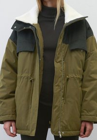 Olivgrüne isolierte Jacke mit einem schwarzen Schulterpanel, Reißverschluss und Kunstfellkragen. Verfügt über einen verstellbaren Kordelzug und Seitentaschen.