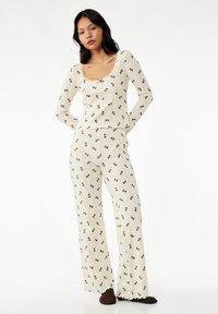 Ribbet pyjamasæt i creme, med firkantet halsudskæring og lange ærmer. Brun sløjfemønster med flagrende bukser og bølgede kanter.