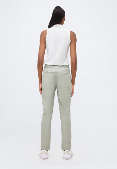 Femme debout, dos à la caméra, portant un haut blanc sans manches, un pantalon gris clair et des baskets blanches sur fond uni.