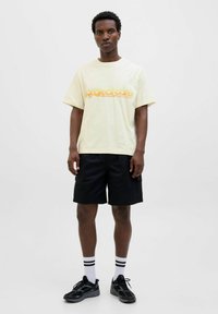 Lichtgele katoenen t-shirt met oranje grafische tekst, gecombineerd met zwarte cargoshorts en zwarte sportschoenen. Witte sokken met zwarte strepen.