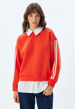 Felpa rossa con strisce bianche sulle maniche, dotata di un colletto bianco sotto, vestibilità comoda e morbida al tatto. Indossata con jeans blu scuro.