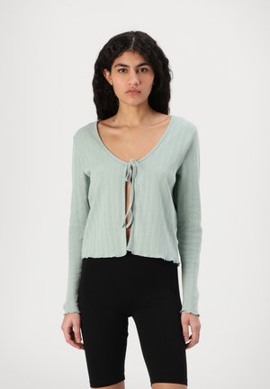 Light green ribbed cardigan with a tie closure, cropped hem, and long sleeves; worn over black bike shorts. Textured fabric, relaxed fit.