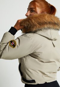 Bežs pūkais jaku ar kažokādas oderi kapuci, melniem adītiem piedurknēm un logotipa uzlīmi uz piedurknes. Iezīmē izliekta apakšdaļa un sānu atvērumi.