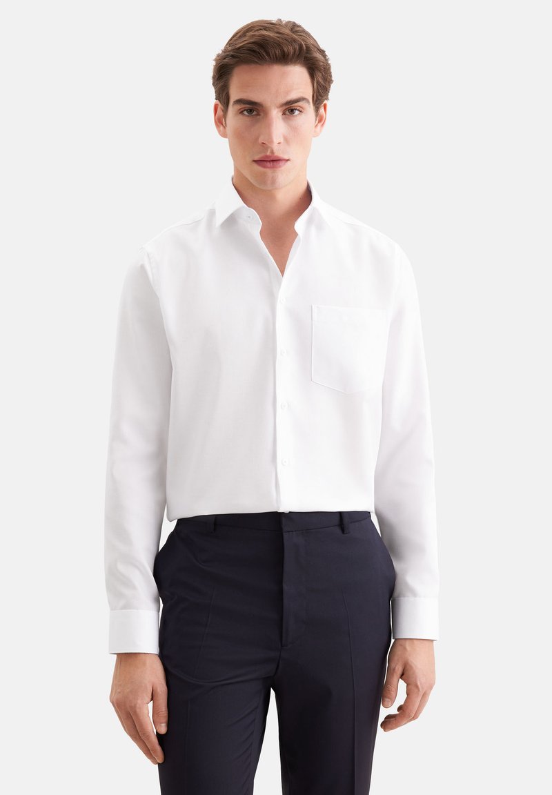 Jeune homme aux cheveux bruns portant une chemise blanche boutonnée avec une poche poitrine et un pantalon bleu marine foncé, debout devant un fond uni.