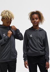 Zwei Kinder tragen dunkelgraue Kapuzenjacken mit Hummel-Logos; eines zieht den Reißverschluss hoch, während das andere entspannt mit den Händen nach vorne schaut.