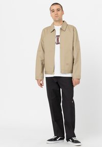 Beige canvasjakke med krage, glidelås og sidelommer, båret over en hvit T-skjorte med burgunder skrift, sammen med svarte bukser og joggesko.