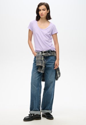 Młoda kobieta stoi z rękami w kieszeniach, ubrana w liliowy t-shirt, szerokie, podwinięte niebieskie dżinsy, czarne buty i koszulę w kratę zawiązaną w talii.