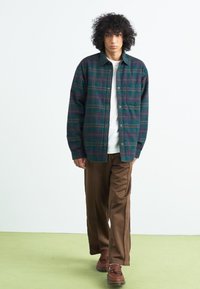 Sötétzöld és tengerészkék kockás flaneling, gombos zárással és mellzsebbel, fehér póló fölött viselve, barna, bő szárú nadrággal párosítva.