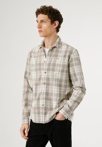 Camicia a quadri in tonalità beige, verde e marrone. Presenta una vestibilità rilassata, maniche lunghe e bottoni in legno. Piccolo logo sul petto.