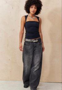 Μαύρο ρουχιέ τανκ τοπ συνδυασμένο με χαλαρό γκρι τζιν παντελόνι, ντυμένο με μια ζώνη με χάντρες και μαύρα παπούτσια, σε φόντο από ανοιχτόχρωμο ξύλο.