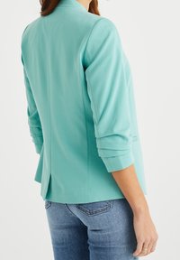 Un blazer verde menta con un corte relajado, mangas de tres cuartos y una abertura en la parte posterior, combinado con unos jeans de mezclilla azul. Textura de tela suave.