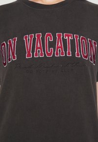 Ciemnoszara koszula z dużym, czerwonym i białym haftowanym napisem "NA WAKACJACH" oraz mniejszym czarnym tekstem "KLUB NIC ROBIENIA" poniżej.