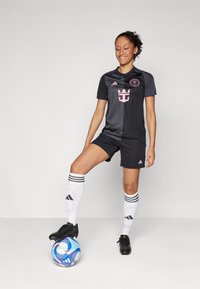 Black soccer jersey with pink accents and logo, paired with black shorts, white striped socks, and black cleats. Blue and white soccer ball.