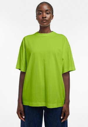 Femme à la peau foncée portant un T-shirt ample vert vif à manches courtes et un pantalon bleu foncé, debout devant un fond blanc uni.