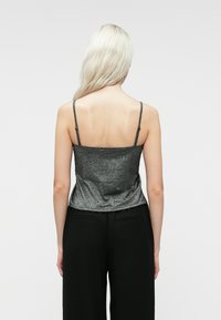 Γυναίκα με μακριά ξανθά μαλλιά, φορώντας μια γυαλιστερή σκούρα γκρι τοπ με λεπτές τιράντες και ψηλόμεσα μαύρα παντελόνια, απεικονισμένη από πίσω.