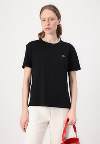 T-shirt en coton noir à manches courtes, col rond et petit logo doré sur la poitrine. Porté avec un pantalon blanc et un sac à main rouge.