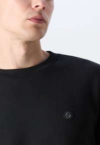 Black crew neck sweatshirt made of smooth fabric, featuring a small round logo patch on the left chest area.