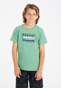 Groene katoenen T-shirt met een rechthoekige grafiek met de tekst "PROTEST" en abstracte strepen, in combinatie met zwarte gebloemde shorts.