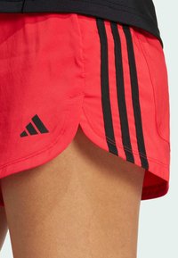 Shorts de sport rouges avec un design à trois rayures noires, arborant un logo noir. Le tissu semble léger et lisse, avec un ourlet arrondi.