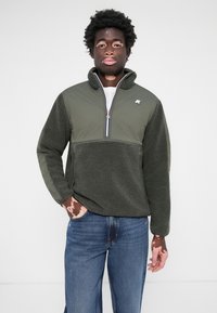 Dark green pullover made of textured fleece with a half-zip collar. Features contrasting smooth fabric on the upper part and sleeves.