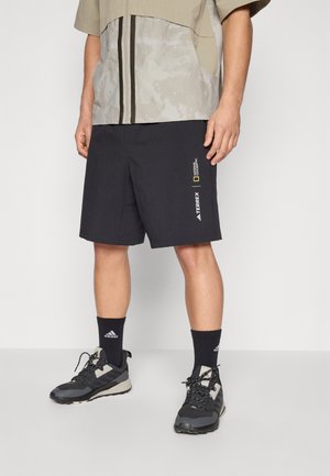 Homme portant un short noir Terrex National Geographic, des chaussettes noires Adidas et des chaussures de sport noires et grises, debout devant un fond uni.