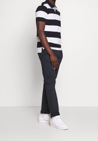 Polo shirt rayé noir et blanc avec un col classique, associé à un pantalon bleu marine et des baskets blanches. Tissu lisse, coupe décontractée.