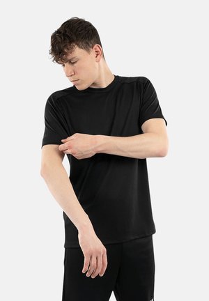 Junger Mann, komplett in Schwarz gekleidet, richtet den Ärmel eines schwarzen kurzärmeligen T-Shirts vor einem einfarbigen hellen Hintergrund.