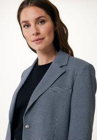 Blazer i svart-och-vitt hundstooth-mönster, med slagkrage, enkel knappstängning och en bröstficka, över en svart topp.