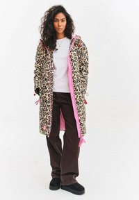 Dlouhý kabát s leopardím vzorem, růžovým zipem a stahovacími detaily, kombinovaný s bílou košilí a hnědými kalhotami s širokými nohavicemi. Měkká tkanina.