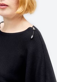 Blond osoba w czarnym prążkowanym swetrze z małym suwakiem na lewym ramieniu na jednolitym białym tle.