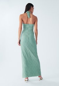 Femme portant une robe maxi dos nu texturée vert clair, debout, tournée de dos, sur un fond blanc uni.
