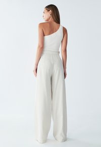 Bela rebrasta majica na jedno rame v kombinaciji s svetlobezž pancaricami širokega kroja, ki se ponašajo z gladko teksturo in minimalističnim dizajnom.