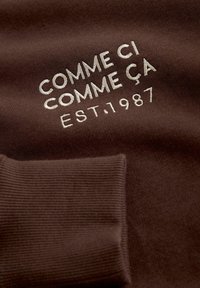 Brauner Sweatshirt mit gerippten Bündchen und weiß gesticktem Text: "COMME CI COMME ÇA EST. 1987." Weicher Stoff mit leichter Struktur.