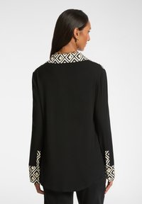 Frau, die sich abwendet und eine schwarze Bluse mit langen Ärmeln und einem geometrisch gemusterten Kragen sowie Manschetten trägt, kombiniert mit schwarzen Hosen und goldener Schmuck.